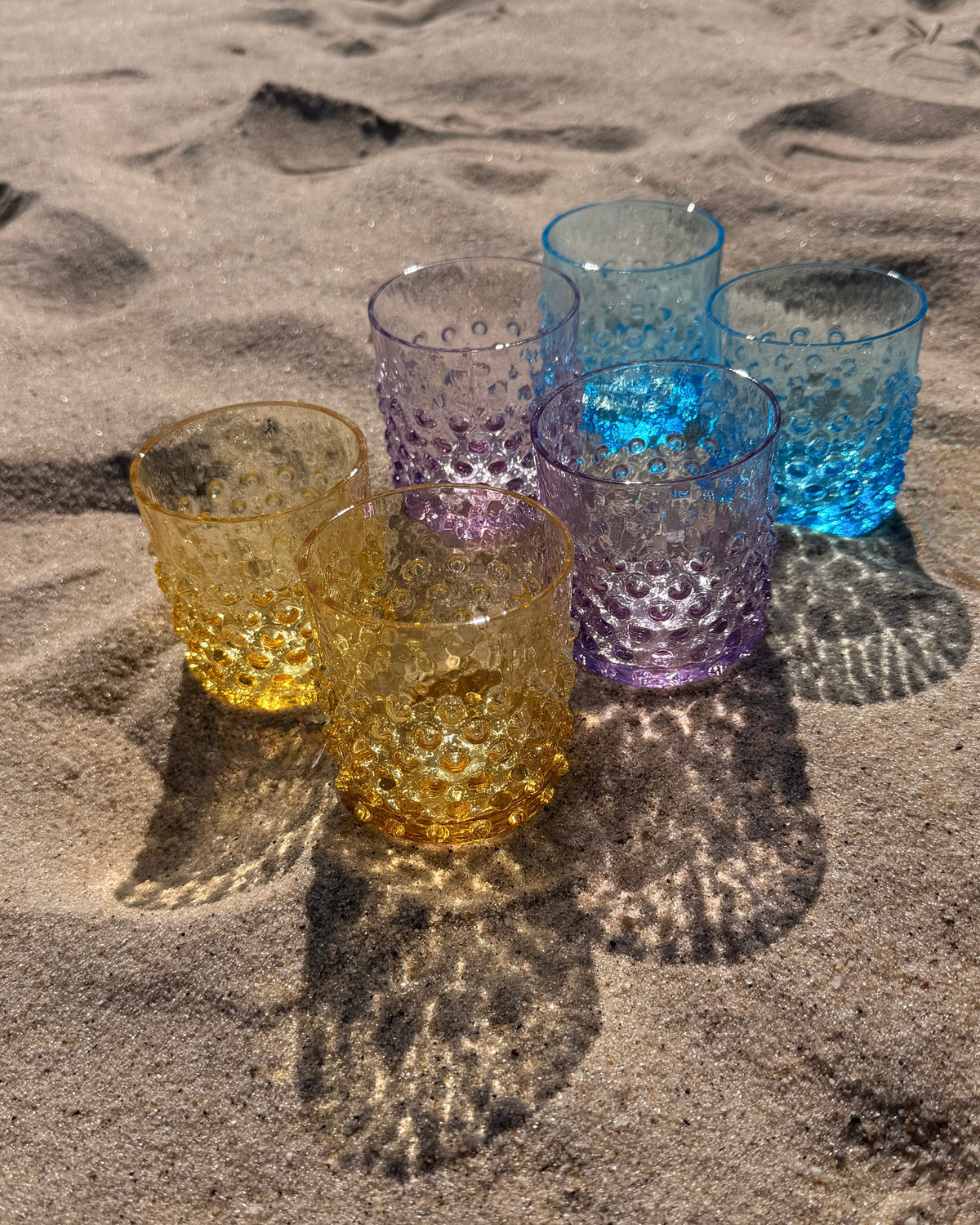 Five textured glass tumblers in Citrine, Lilac, and Azur are arranged on sandy ground outdoors, casting shadows on the sand.