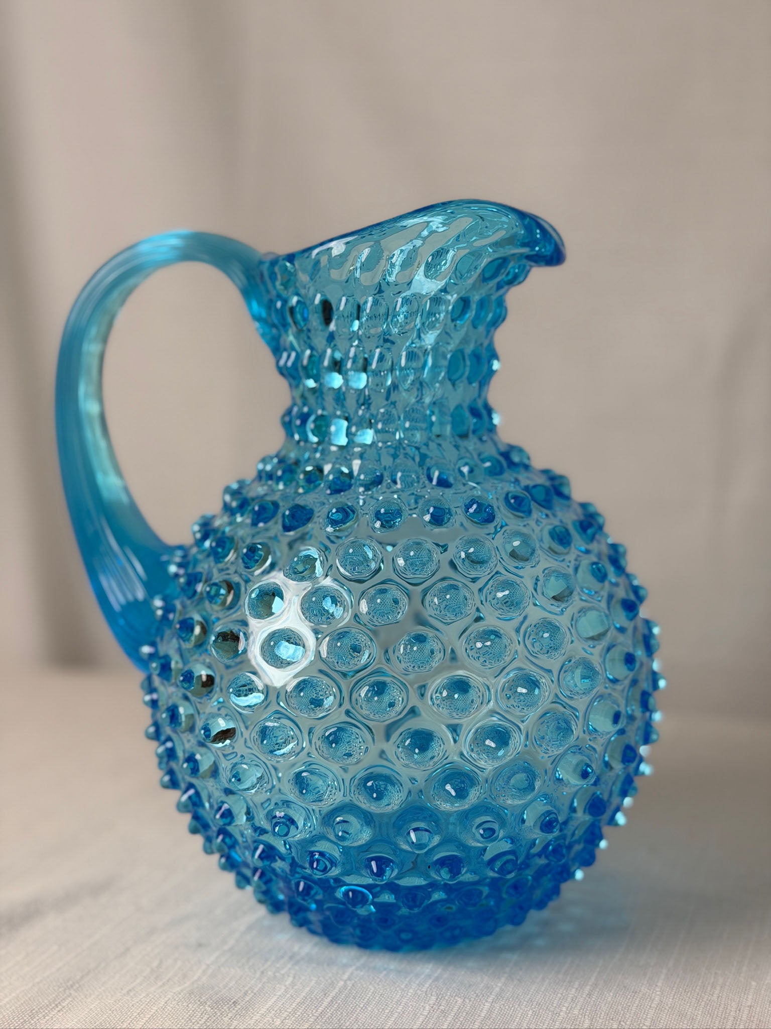A vintage-style Azure hobnail glass pitcher with a textured, bubble-covered surface and a curved handle, set against a neutral beige fabric background.