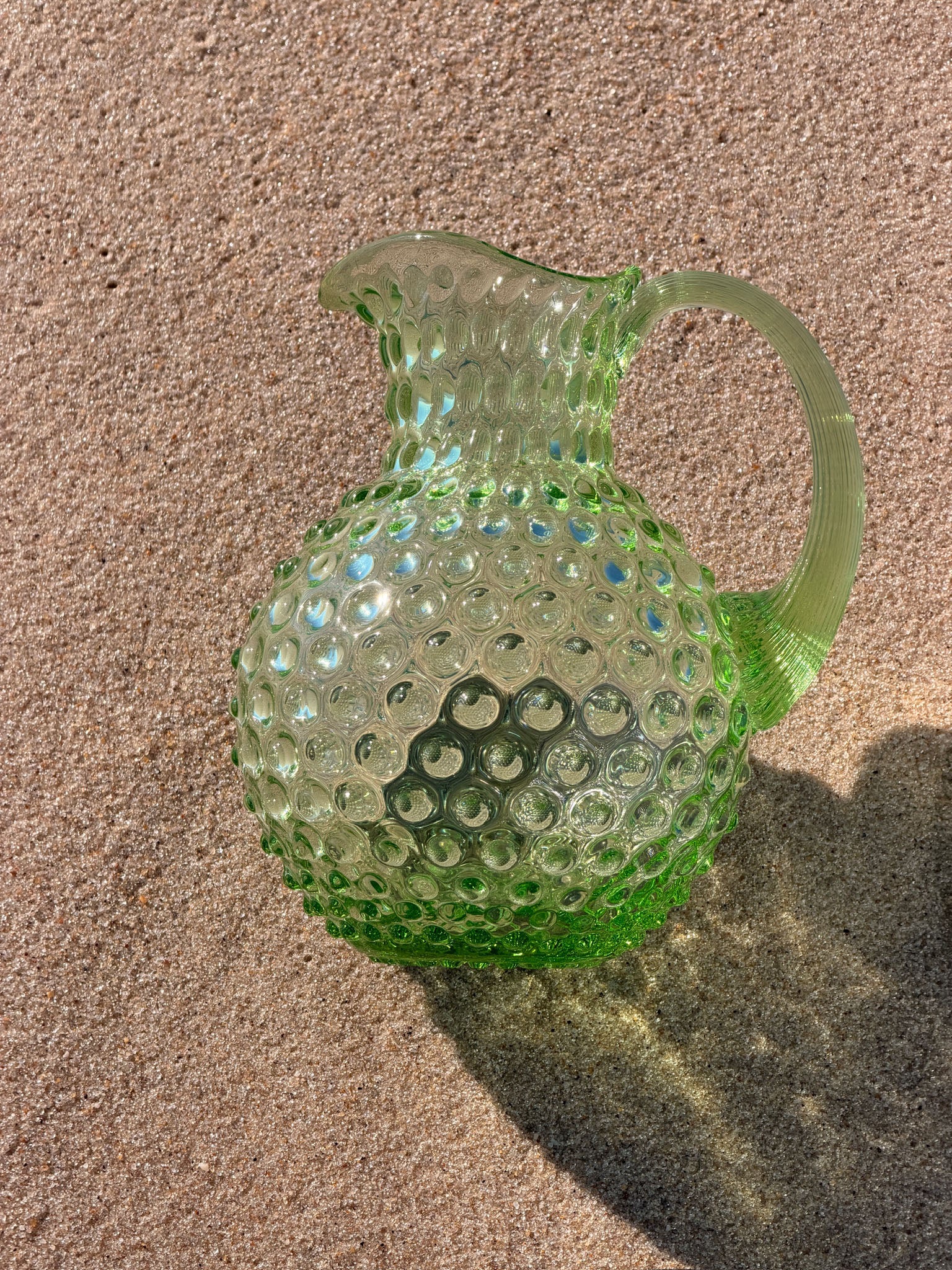 A light green hobnail glass pitcher with a rounded body, spout, and handle, resting on a sandy surface with a shadow cast to the right.