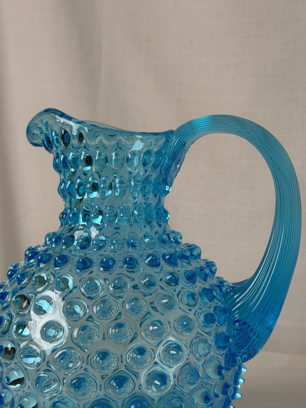 A close-up of a translucent Azure glass pitcher with a textured hobnail pattern and a curved handle, set against a neutral beige fabric background.