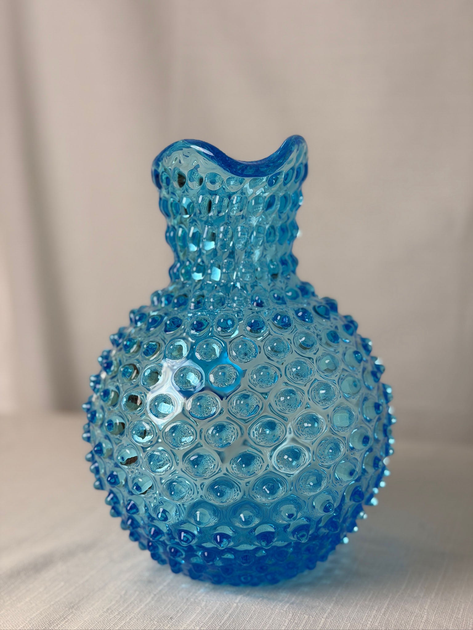 A small hobnail-textured glass vase in Azure, featuring a rounded body and a narrow, flared neck, set against a neutral light gray background.